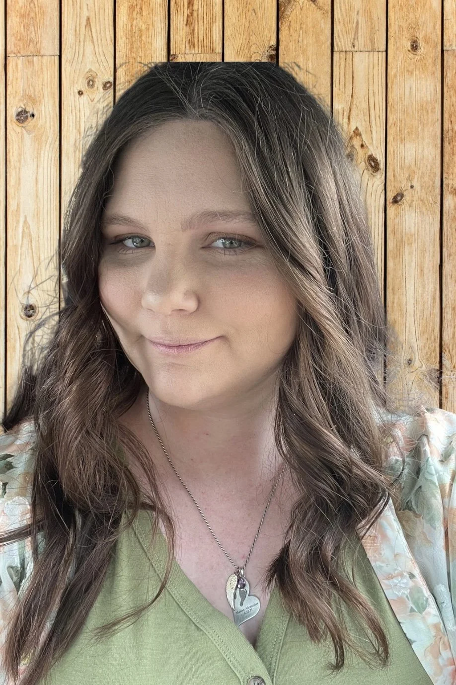 A woman with long wavy brown hair and blue eyes smiling slightly, wearing a green top and a floral cardigan in front of a wooden wall.