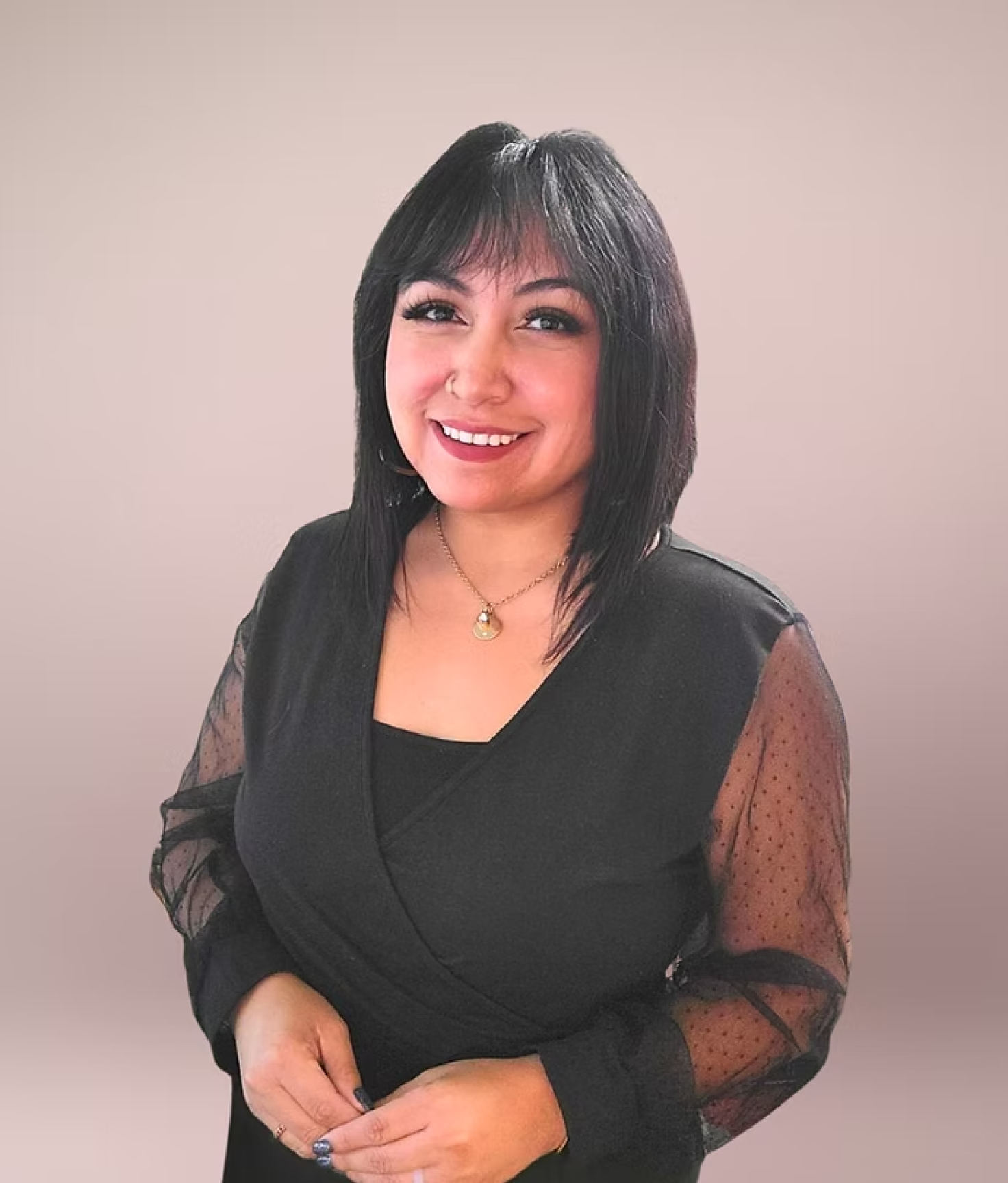 A woman smiling, with black hair and wearing a black top with sheer, polka-dotted sleeves, standing against a plain, light-colored background.