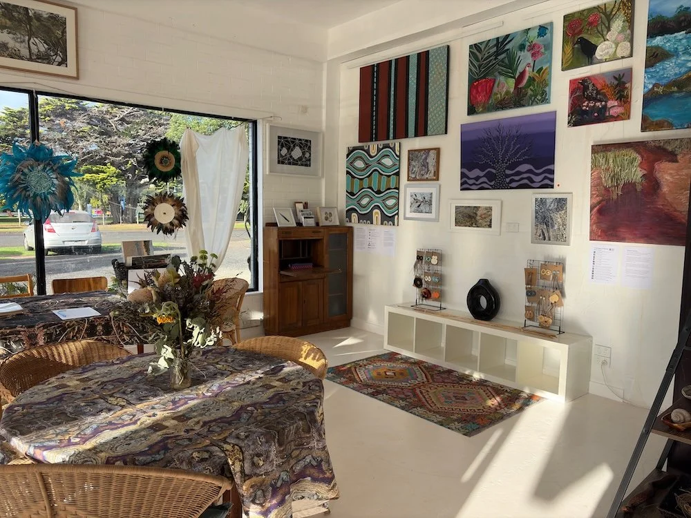 An art gallery interior with colorful paintings on white walls, a windows with outdoor view, a table with a patterned tablecloth and floral arrangement, and various art displays and furniture.