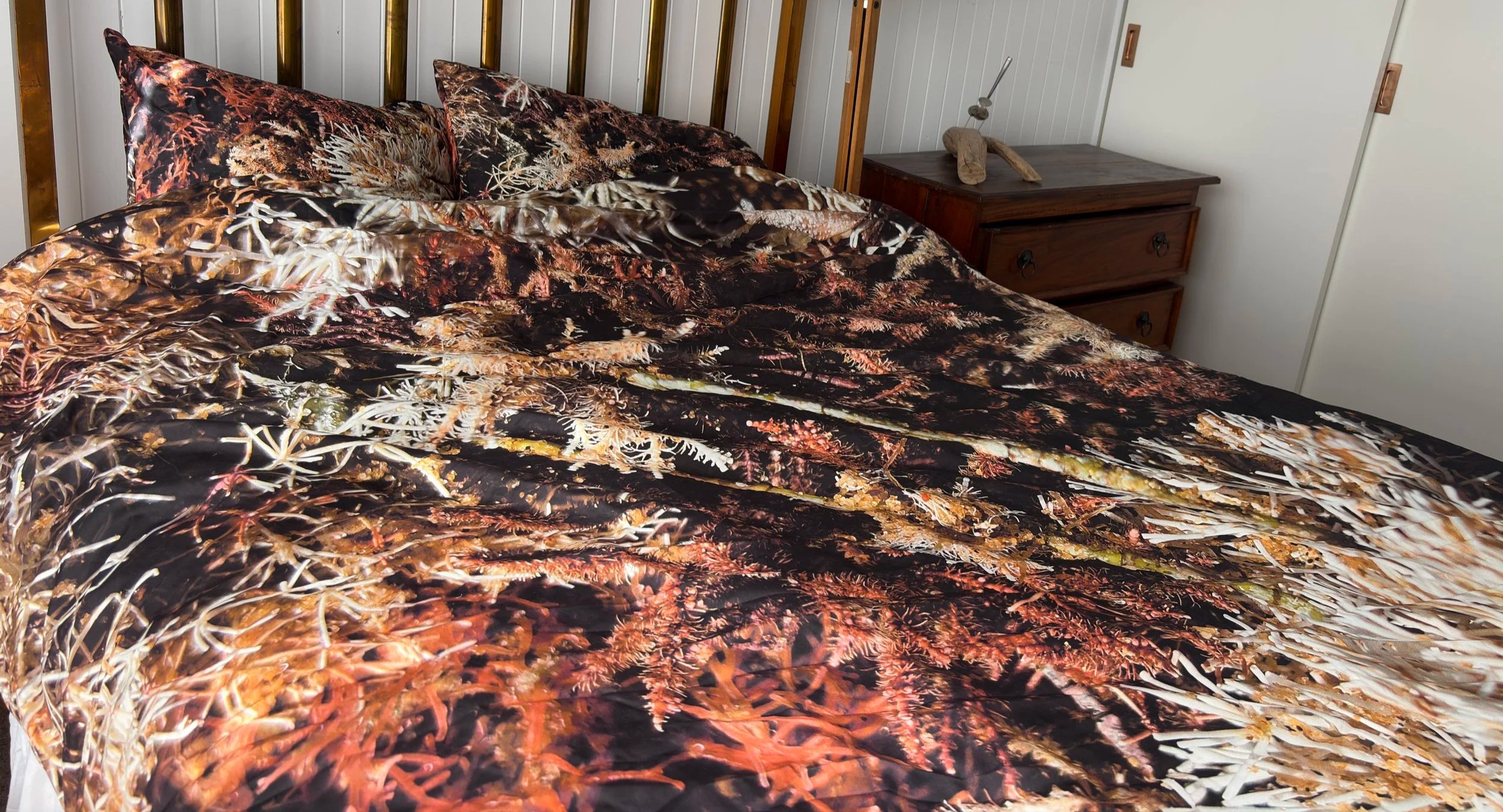A bed with a duvet or doona cover featuring a vivid coastal scene of seaweed and algal forms in rich reds, browns, oranges, creams and white, along with matching pillows, in a bedroom with white paneled walls and a wooden nightstand.