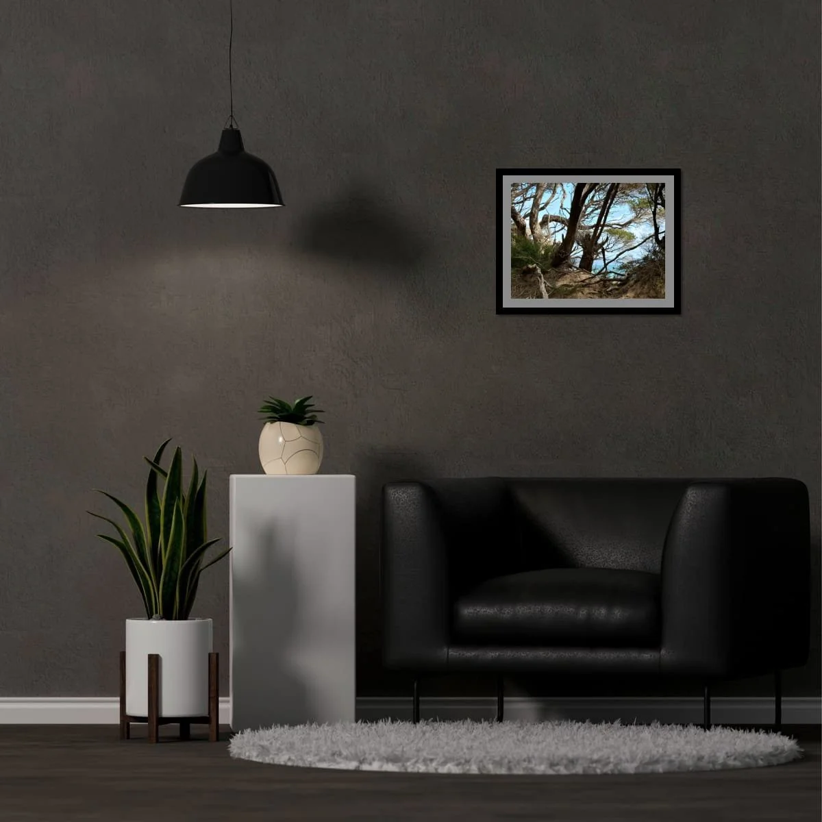 Framed photograph of coastal trees displayed on a dark grey wall above a black armchair, styled with a hanging pendant light and grey plinth
