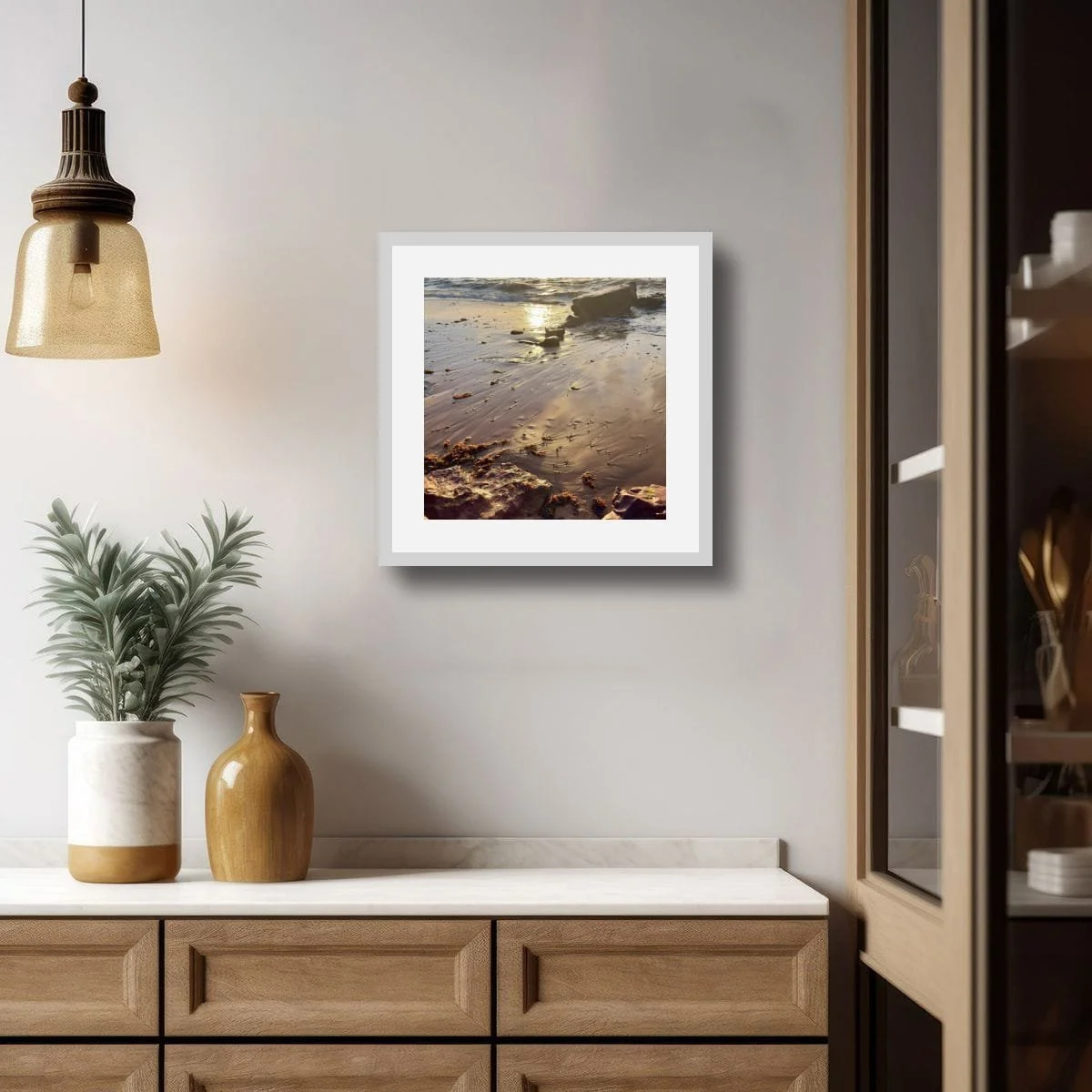 Framed coastal photograph in white mat, hung in warm timber interior with golden pendant light and ceramic vessels.
