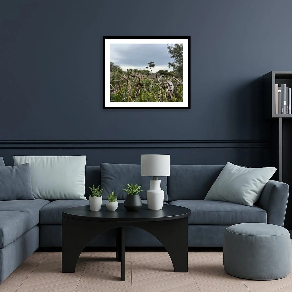 Framed print of Listening Field on a deep navy wall above a blue-grey sofa with pale cushions. Round black coffee table with small plants and lamp in front.