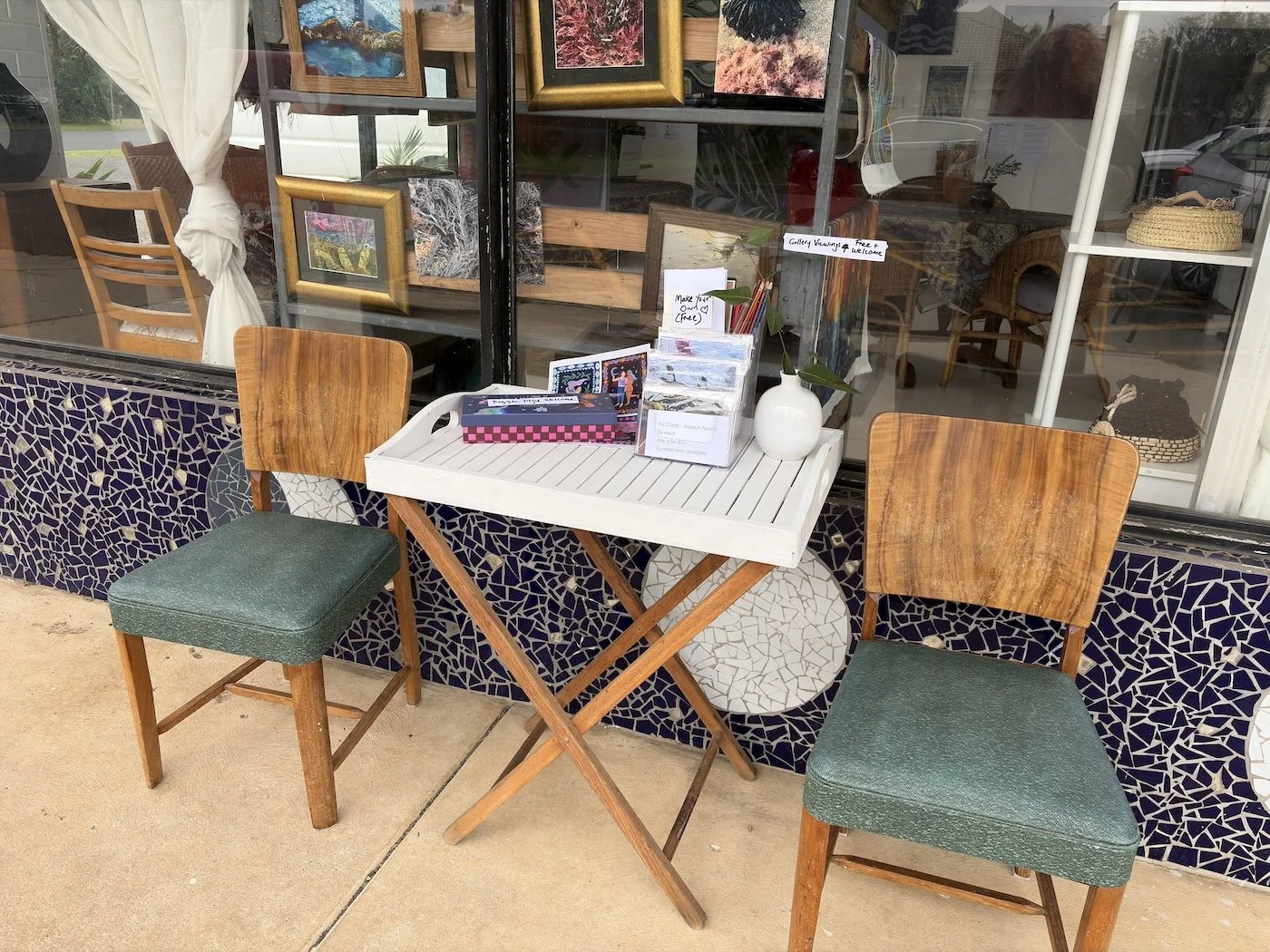 A small outdoor setup with a white folding table flanked by two wooden chairs with art cards and activities. Behind it, a large window displays framed artwork and baskets inside the art gallery.