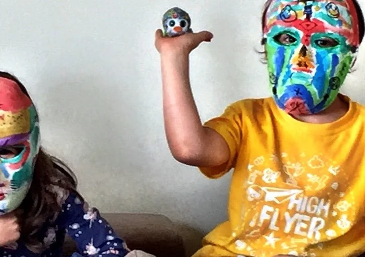 Two children wearing colorful, decorated masks, one holding a small toy and the other raising their arm, sitting in front of a plain wall.