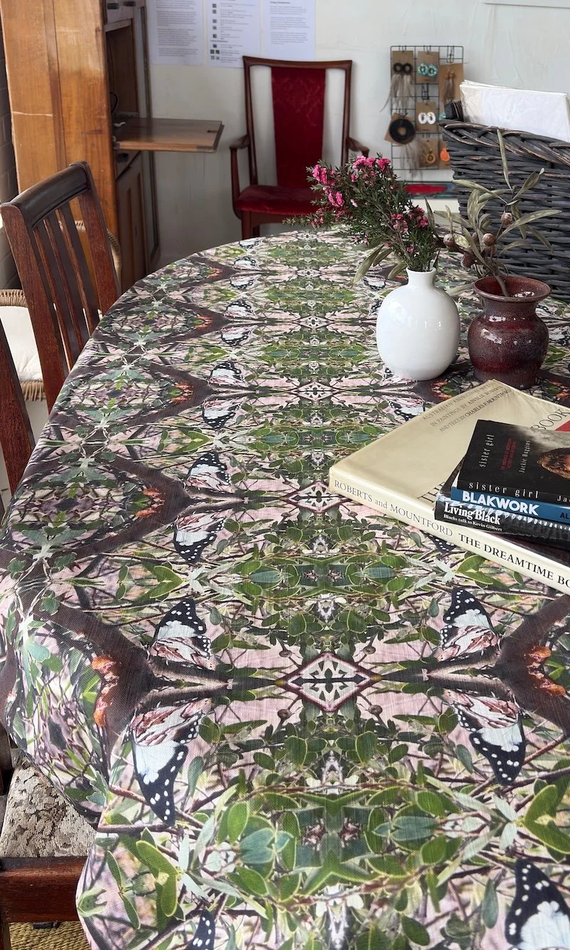 A dining table covered with a butterfly-patterned tablecloth with books, a white vase with pink flowers, and a brown vase with olive branches on top. In the background, there is a red upholstered chair and some papers on the wall.