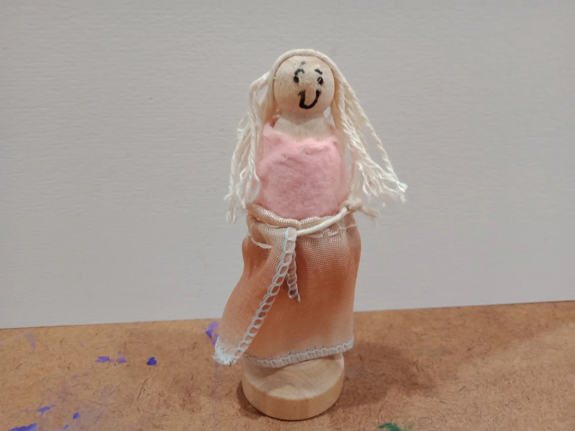Small handcrafted doll with a smiling face, long yarn hair, pink felt top, and brown fabric skirt, standing on a wooden base.
