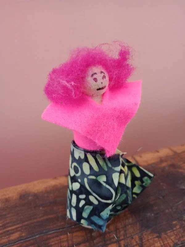 A small wooden puppet with pink felt hair, a smiling face drawn on a round head, wrapped in patterned cloth, standing on a wooden surface against a plain background.