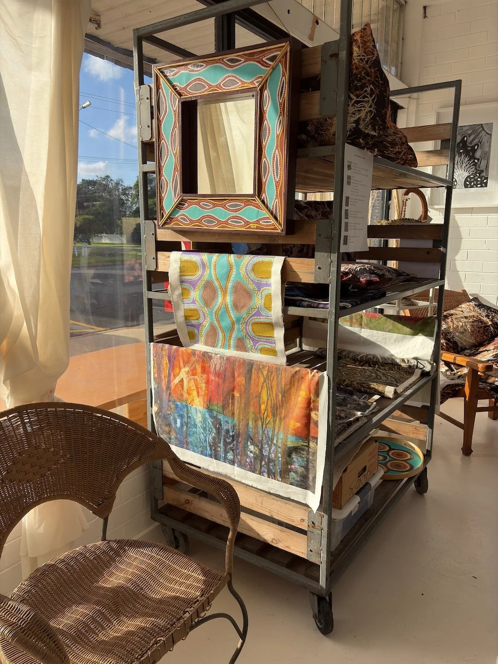 A metal and wood shelving unit displaying colorful textiles, a mirror, and artwork inside a bright room with white brick walls and large windows.
