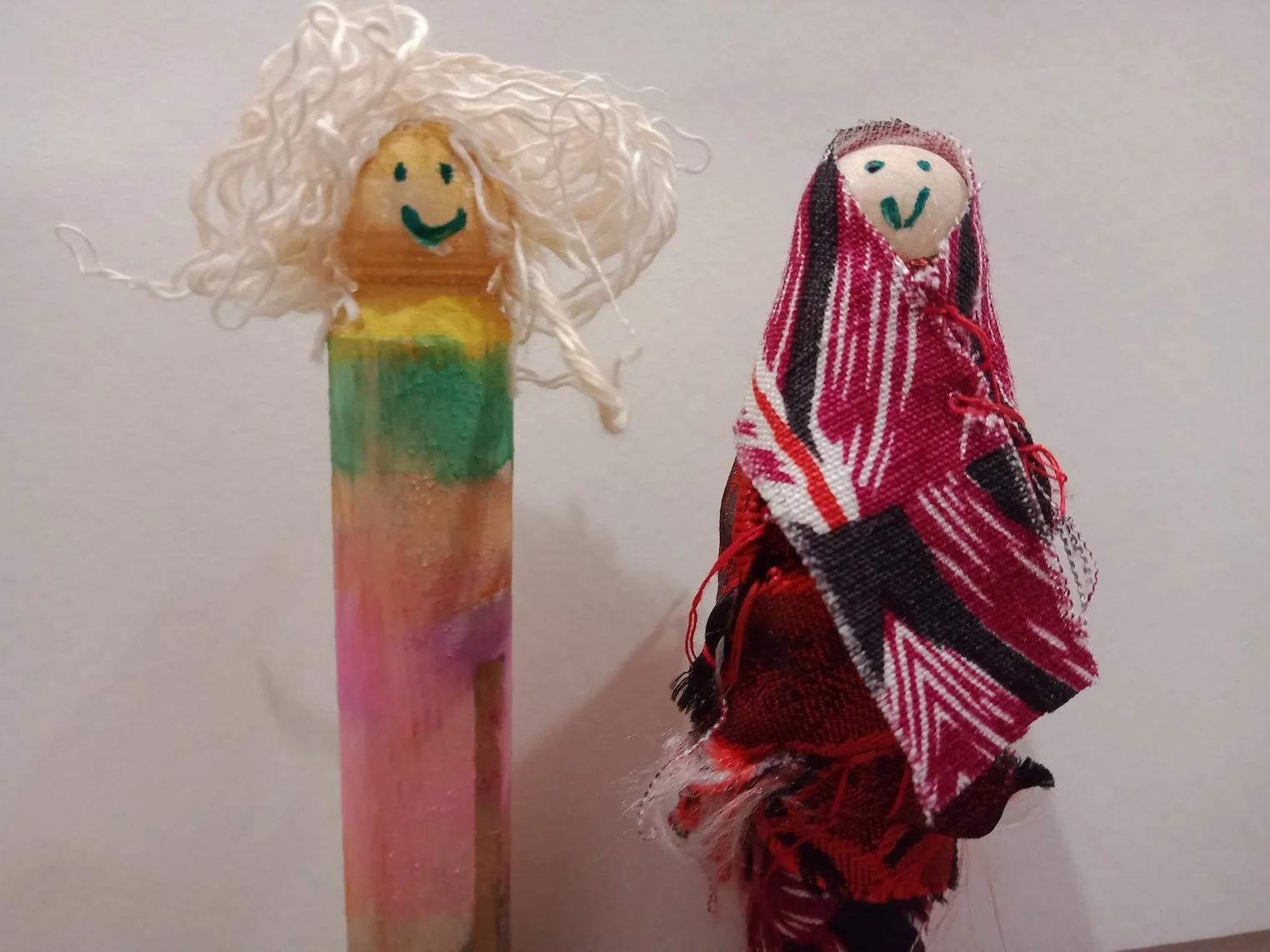 Two wooden dolls, one with a smiley face and white yarn hair, standing next to a small figure wrapped more fully in fabric suggesting muslim clothing.