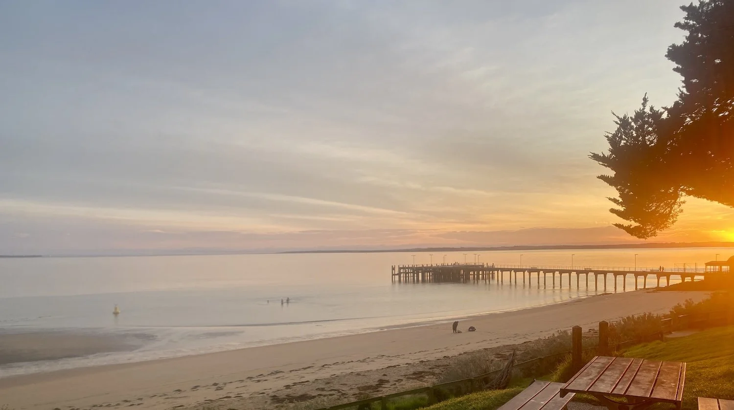 Beach scene at sunset with a pier extending into calm water, a few people on the shoreline, trees on the right, and a picnic table in the foreground.
