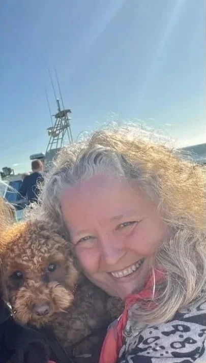 A woman smiling and holding a small, curly-haired brown dog on a boat, with the ocean and a clear blue sky in the background.