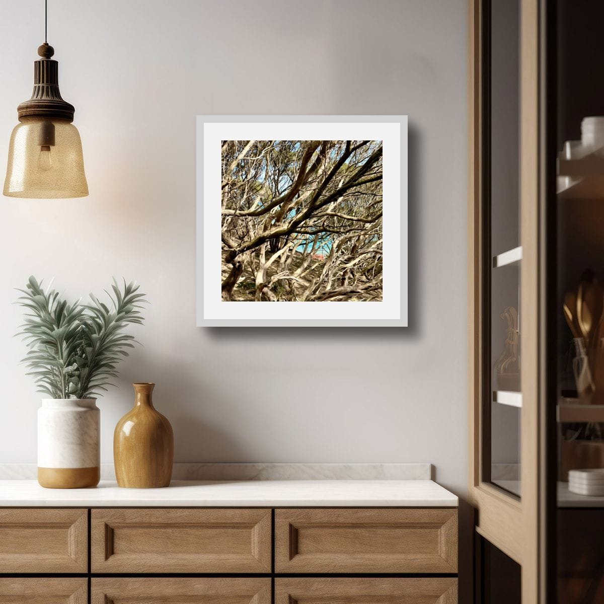 Framed photograph of intertwined coastal tree branches hanging on a soft grey wall above a timber sideboard, styled with a pendant light, plant and ceramic vessels.