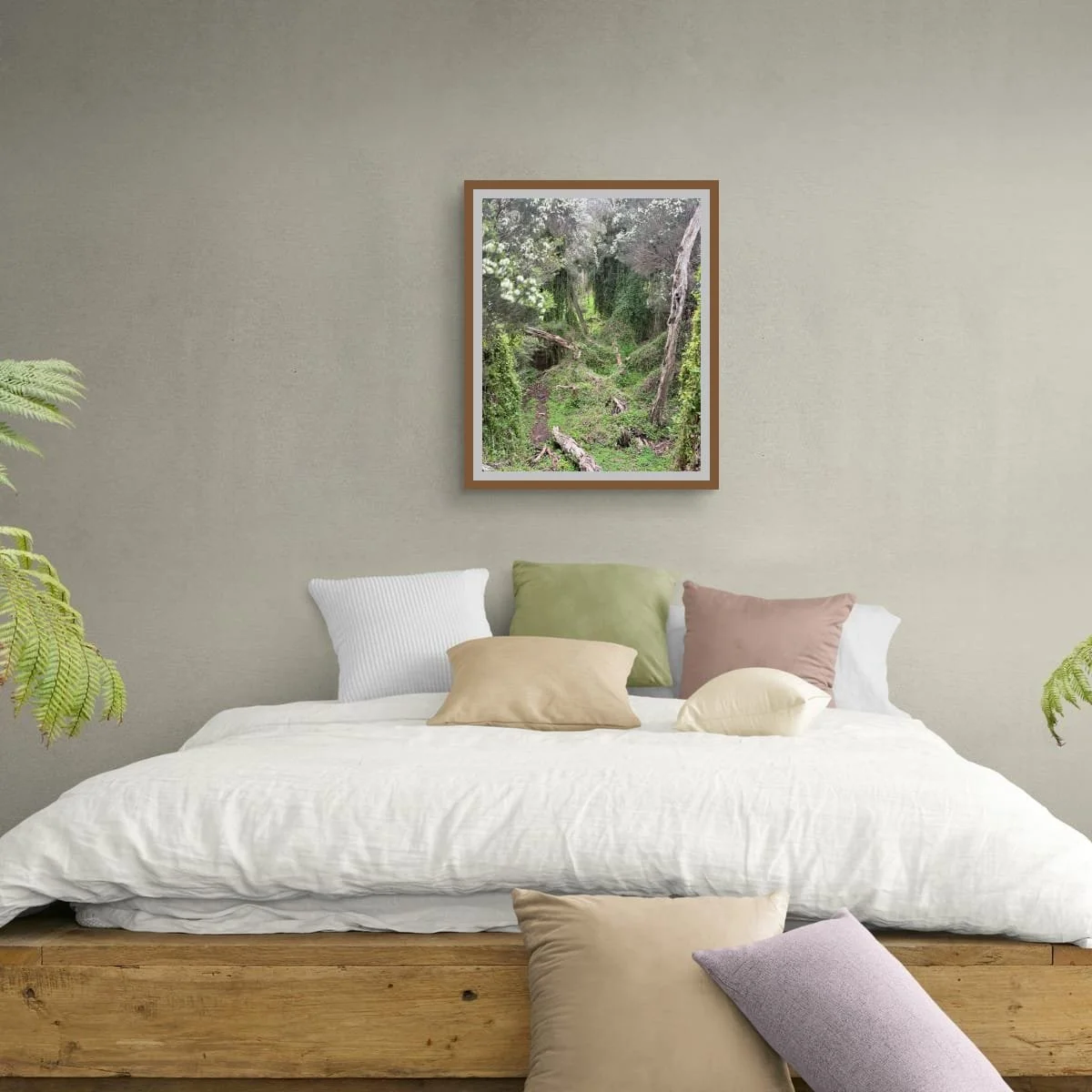Framed print of Understory above a timber bed with white linen and cushions in sage, blush and cream. Two ferns flank the bed. Warm timber frame, grey-green wall.
