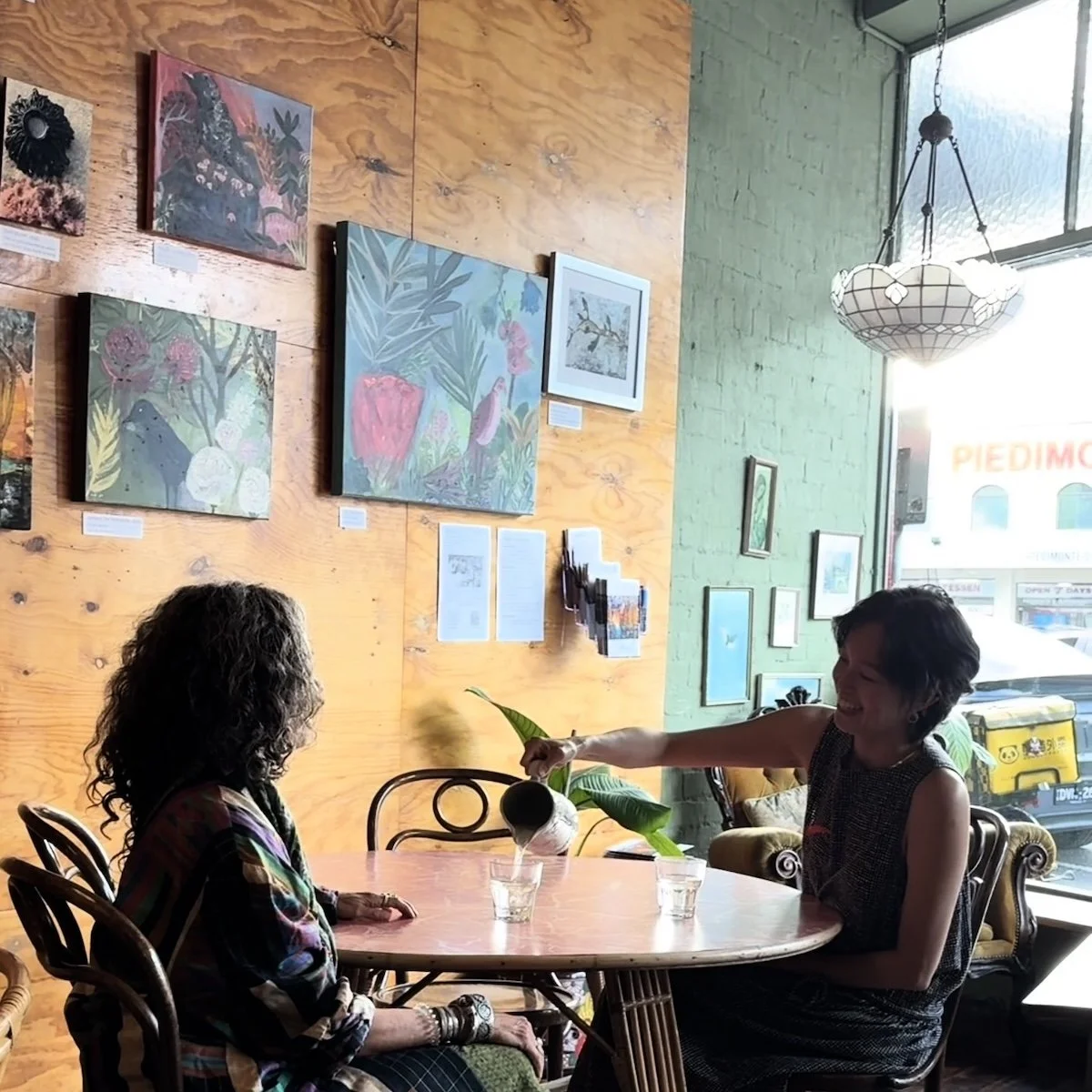 Two women sitting at a round table in a cozy cafe, one pouring a drink for the other. The cafe has wooden and green painted walls decorated with colorful artwork and framed pictures, with a hanging light and a large window letting in natural light.