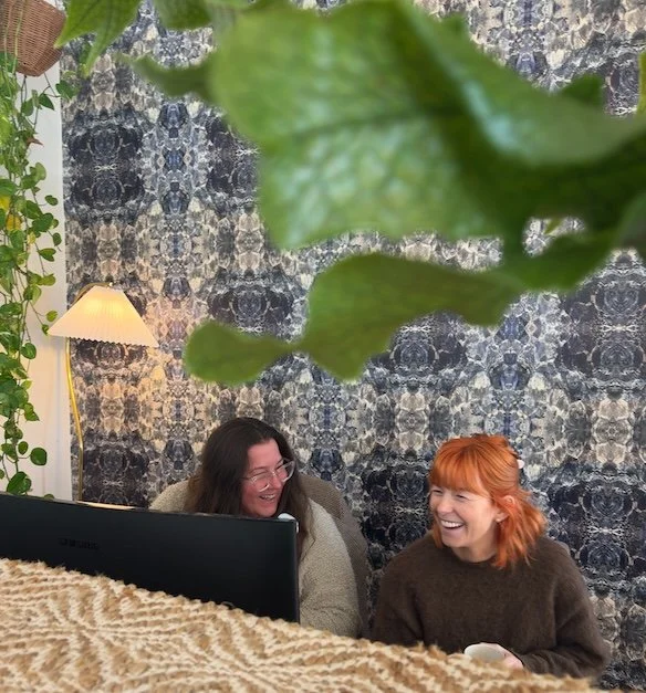 Two women with glasses laughing and talking at a table with a computer in front of them, surrounded by houseplants, with a leafy plant in the foreground and a patterned blue wallpaper on the wall behind them.