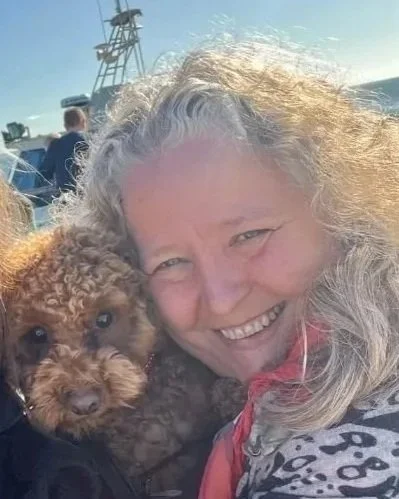 A woman smiling and hugging a small, curly-haired red dog with a boat and blue sky in the background.