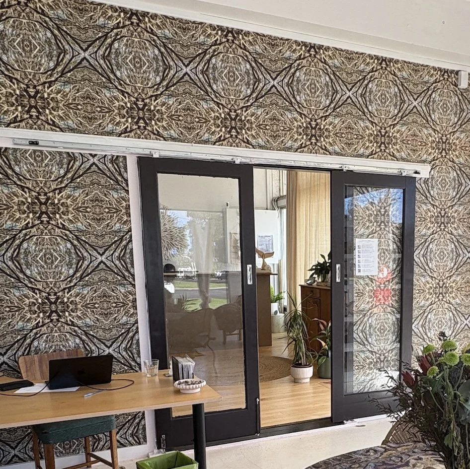 An organic abstract wallpaper repeat in natural greens and blues in a large room with a wooden desk, black laptop, desk supplies, and a wooden chair. The wall is separated by glass sliding doors from another room.