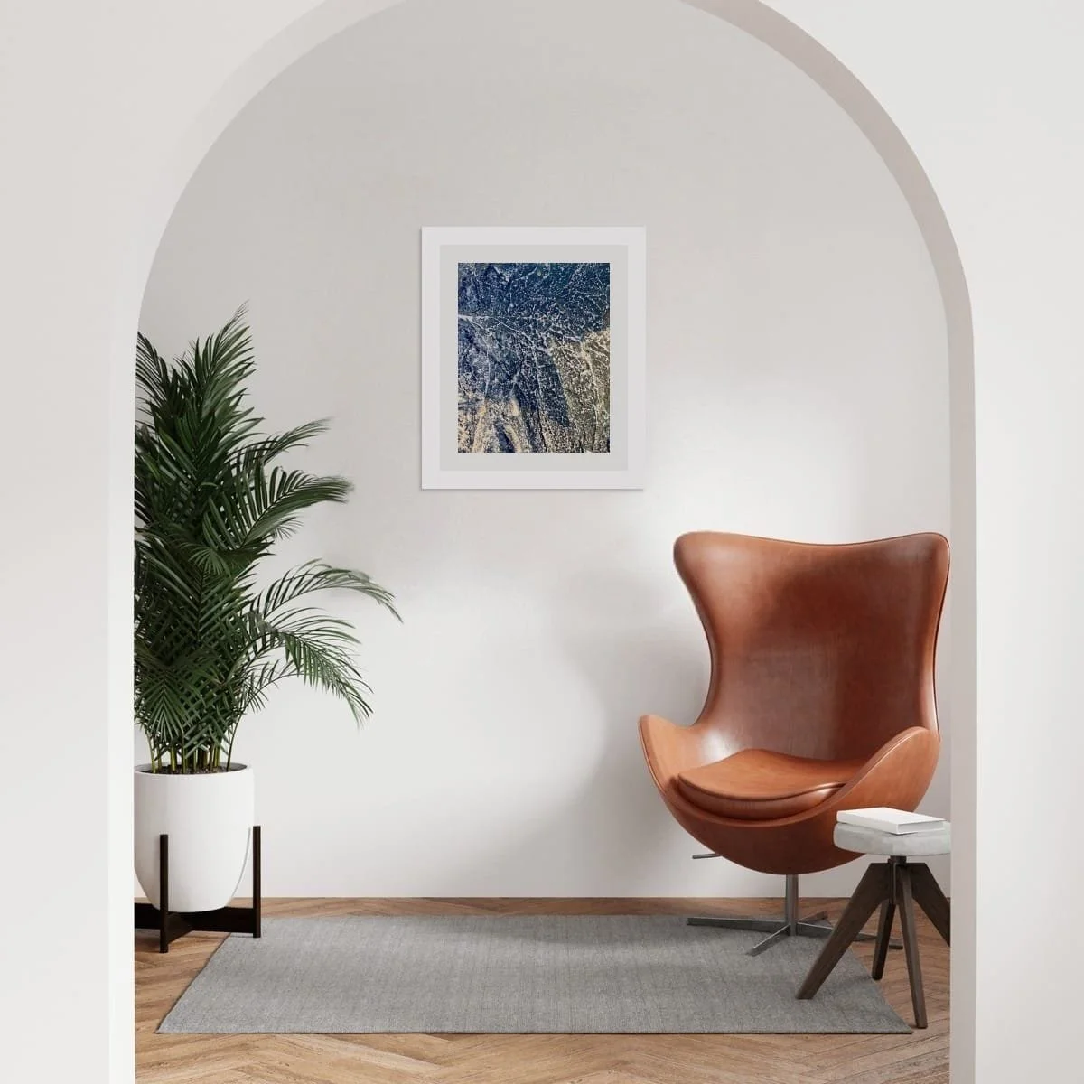 Interior mockup with a framed earthern currents print centred within a curved wall niche, above a leather chair and potted plant,
