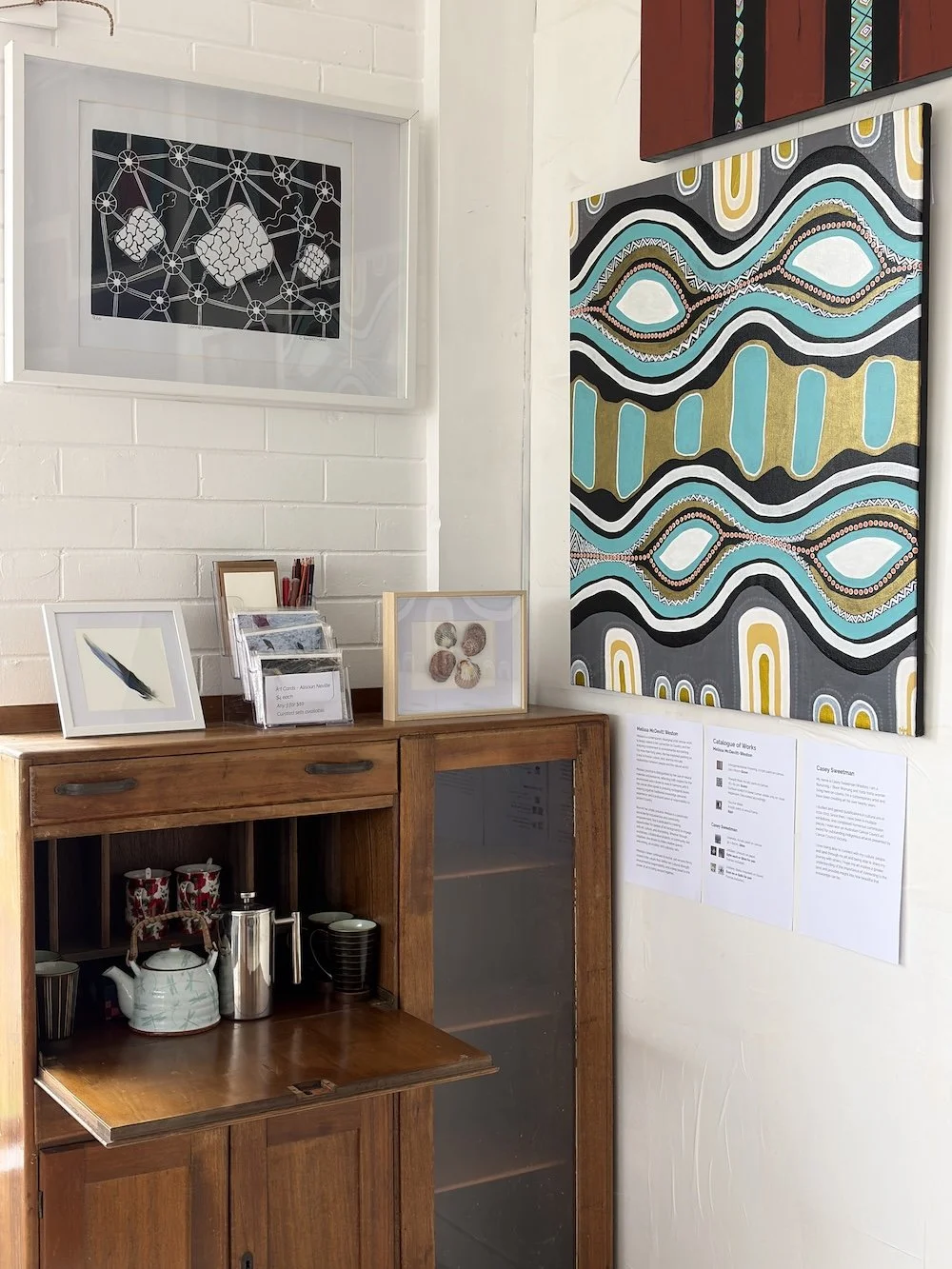 Corner of an art gallery with framed artwork and a wooden cabinet holding cups, teapots, and a coffee plunger.
