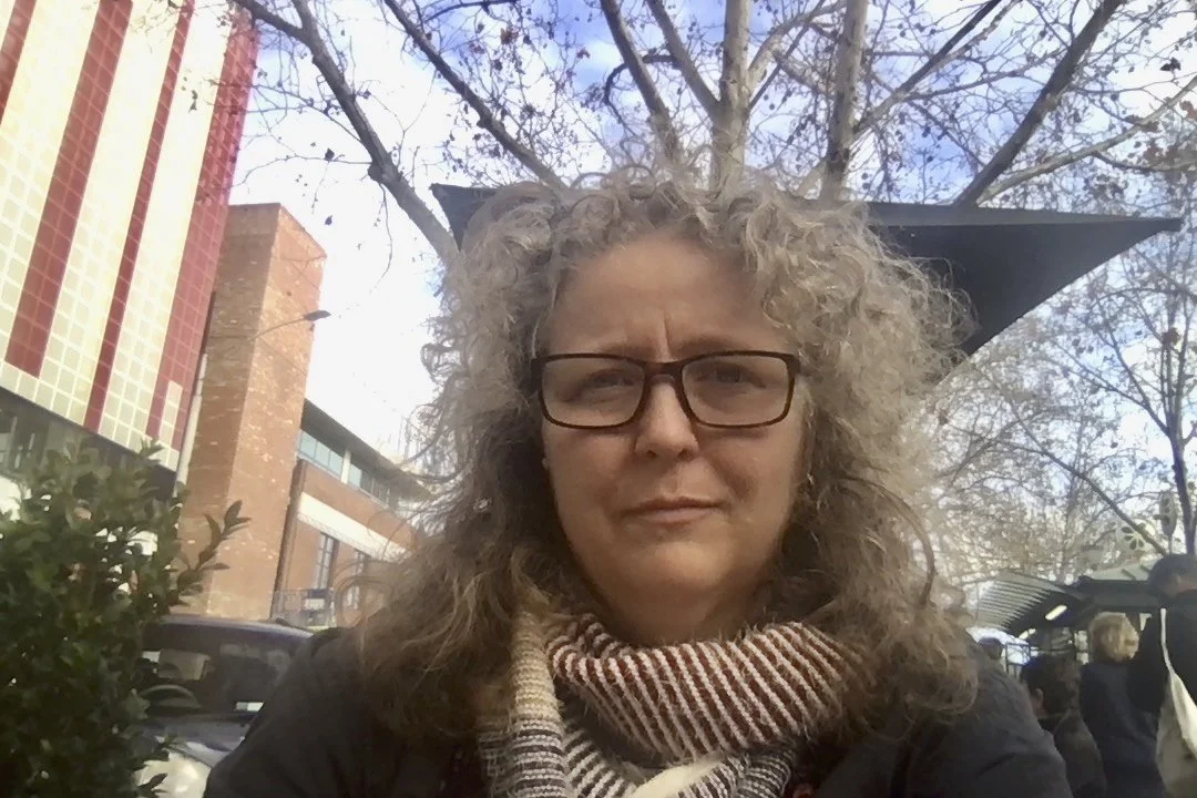 A woman with curly gray hair and glasses wearing a scarf, outdoors with trees and a building in the background.