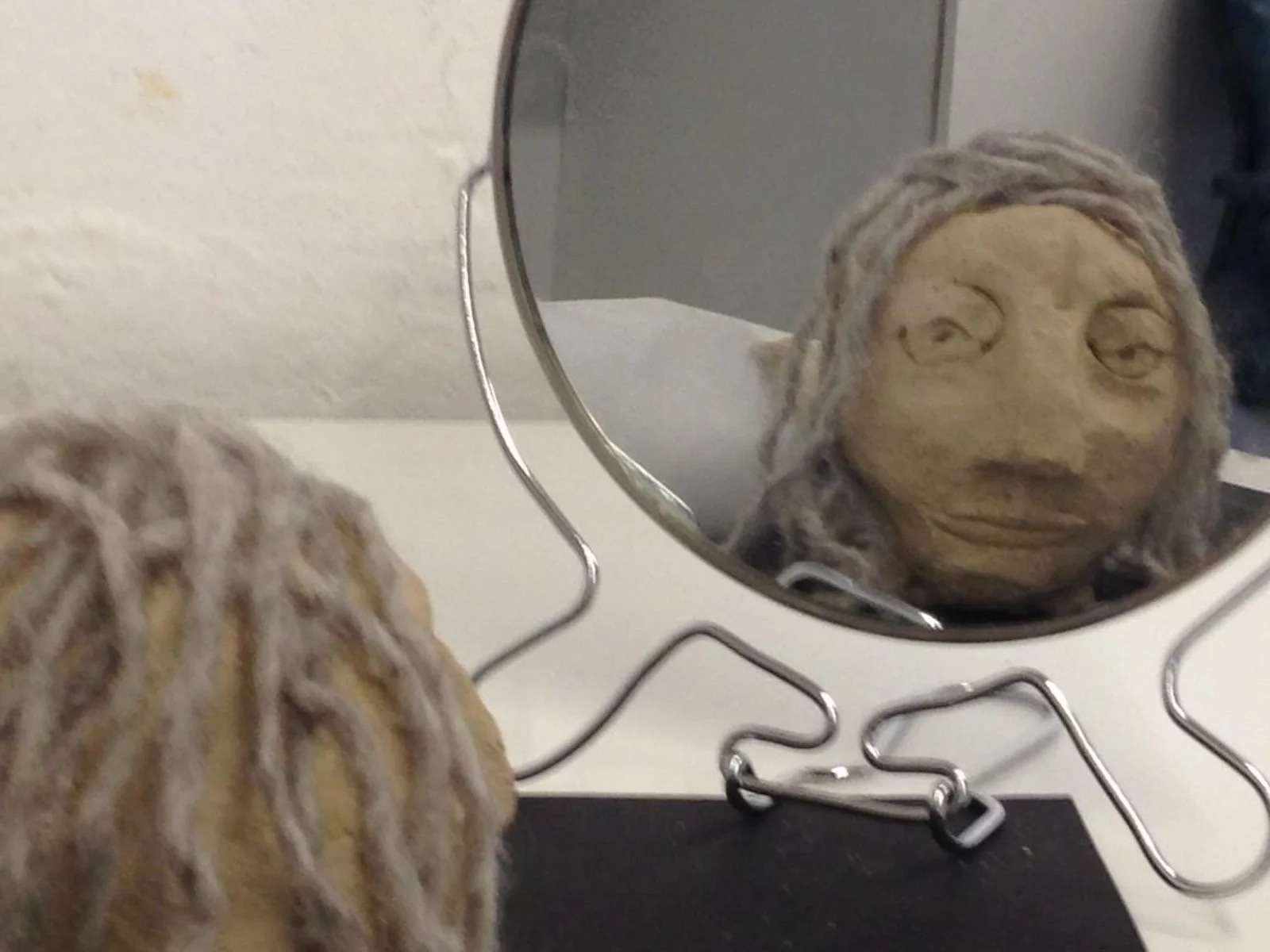 A clay face sculpture with gray hair looking into a mirror.