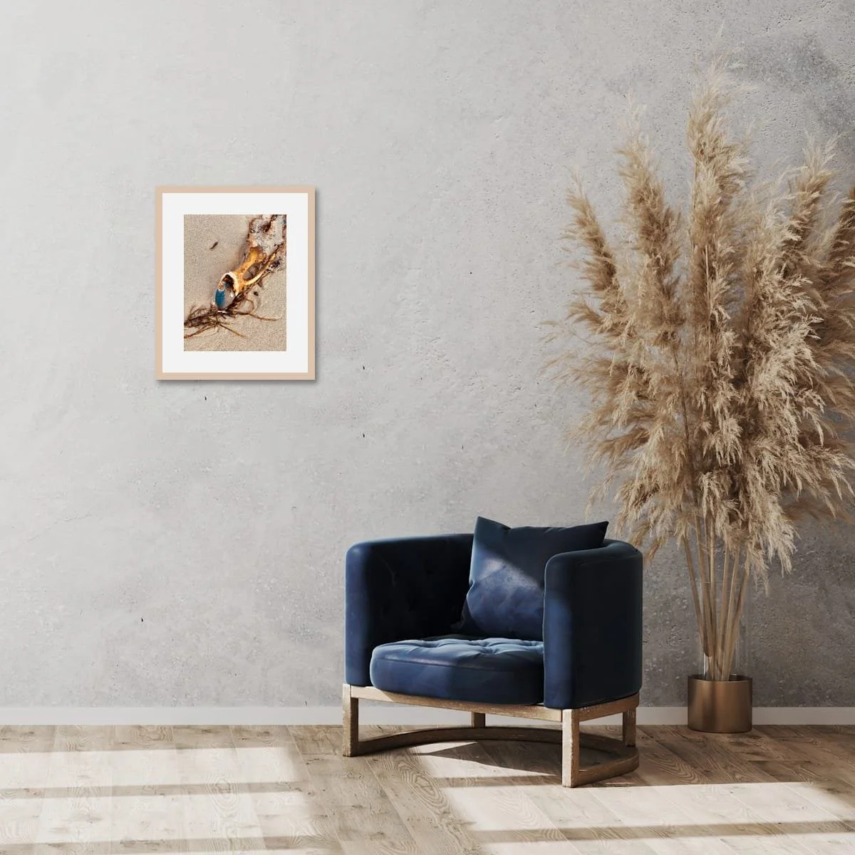 Framed artwork of a kelp holdfast holding a stone, displayed on a pale wall above a blue armchair and dried grasses in a minimal interior.