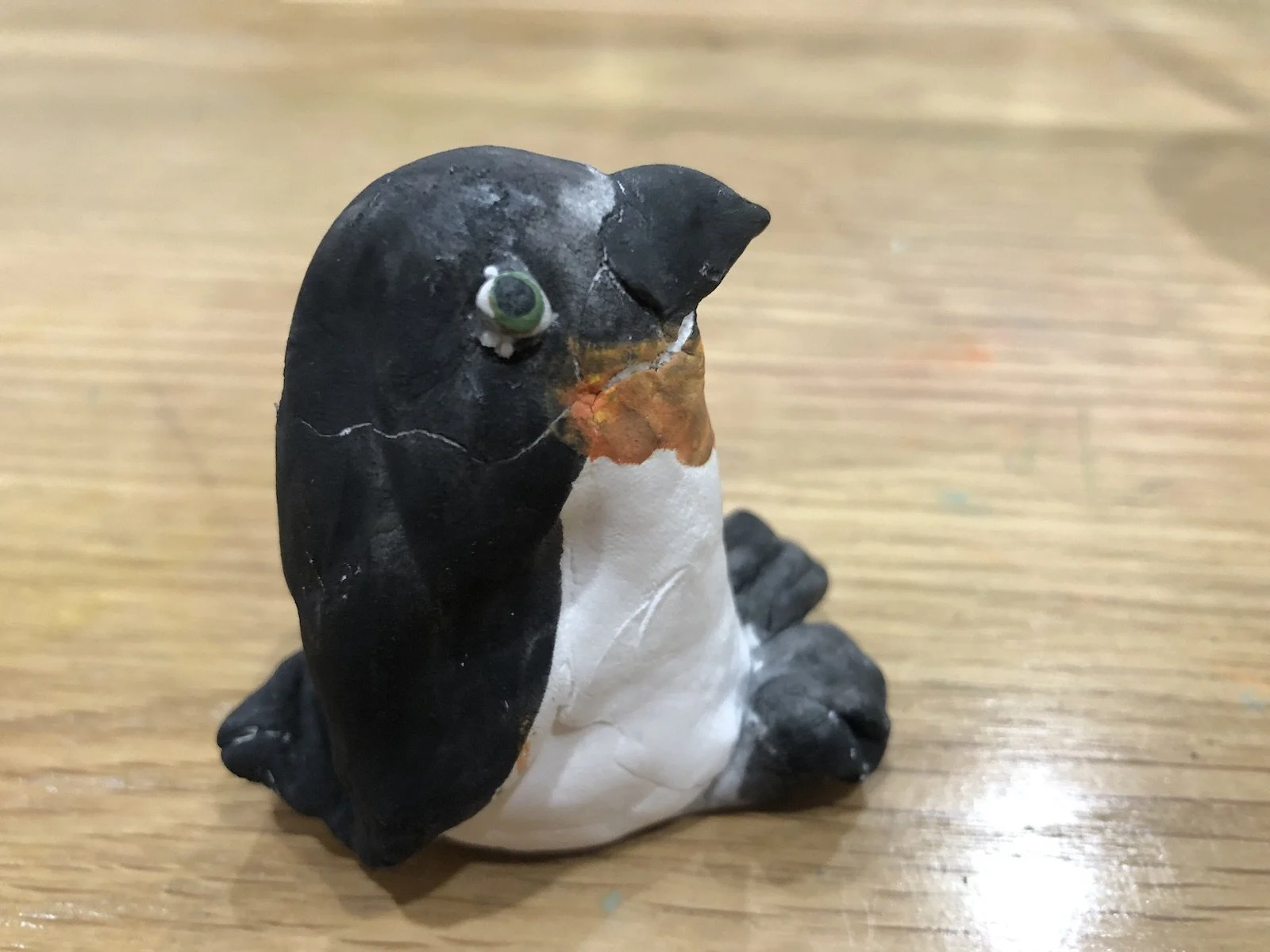 A small penguin figurine with a black body, white belly, orange beak, and green eyes, sitting on a wooden surface.