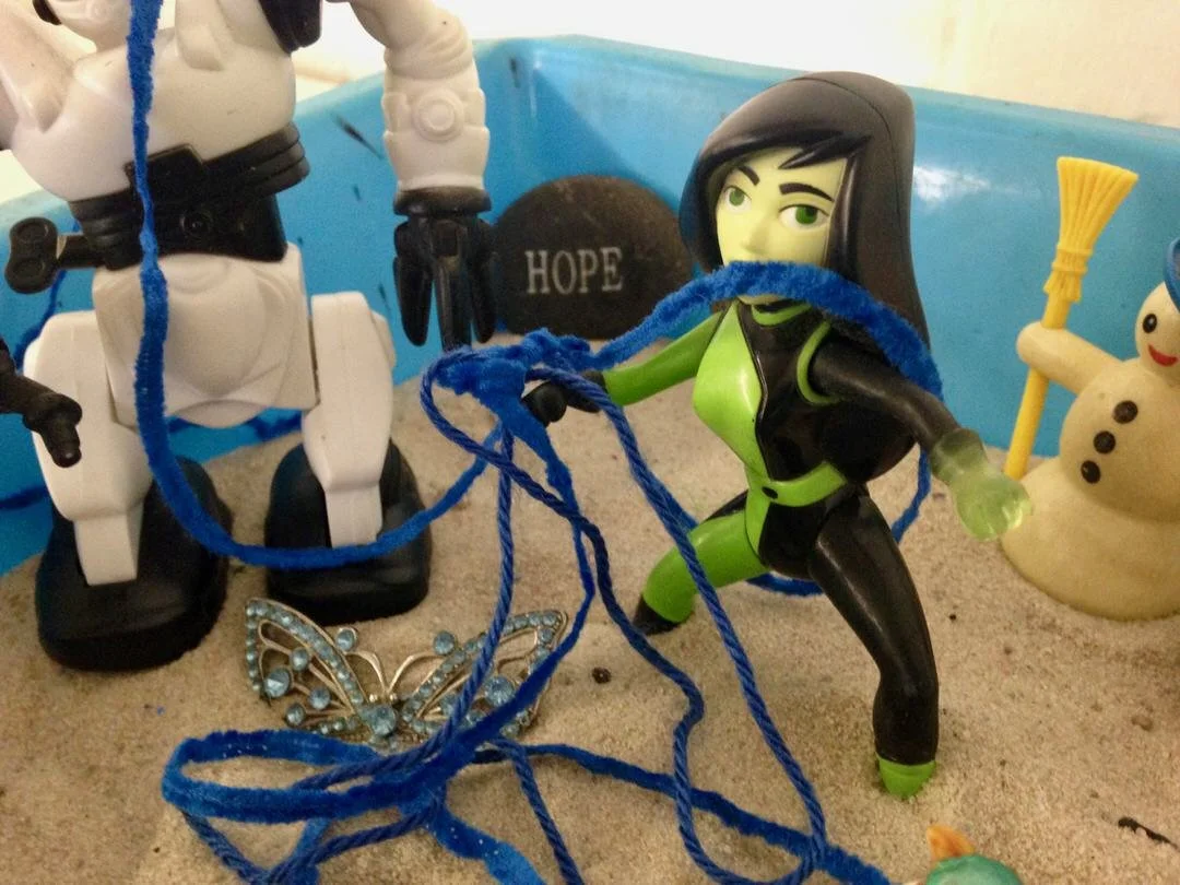 Toy figurines including a female character in black and green with long black hair, an astronaut, a butterfly-shaped decorative item with blue stones, a small snowman, and a rock with the word 'HOPE' written on it, all arranged on sand.