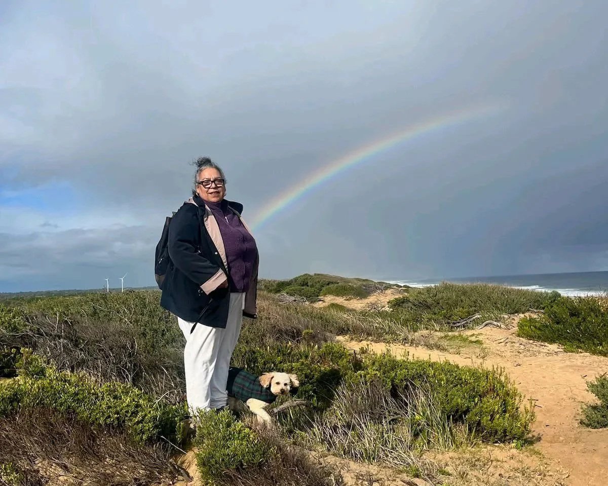 A woman and small dog stand in low lying coastal scrubland in front of a distant ocean and a rainbow in blue sky.