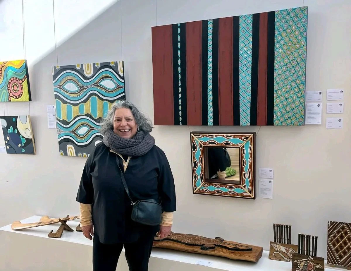 A stands in front of a wall of Aboriginal paintings sitting above cultural works in wood and other materials.