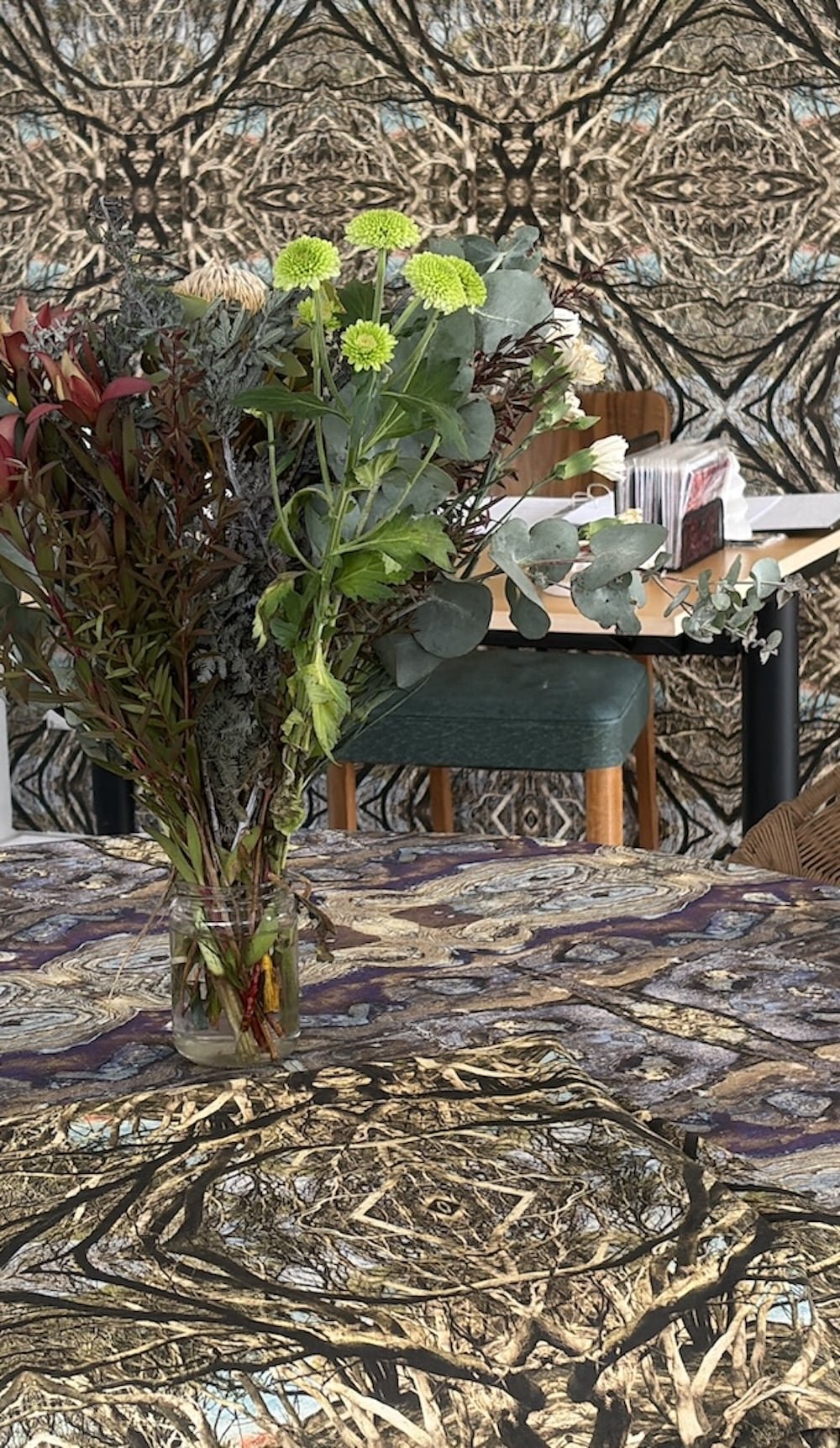 A bouquet of mixed flowers and greenery in a glass vase on a patterned tablecloth, with a background of a wall decorated with a symmetrical, organic abstract wallpaper.