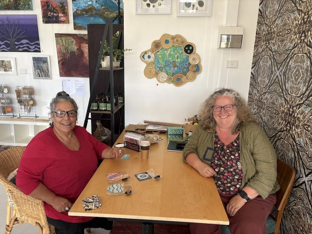 Two women facing the camera sitting either side of a table filled with art supplies and artworks on the wall behind them.