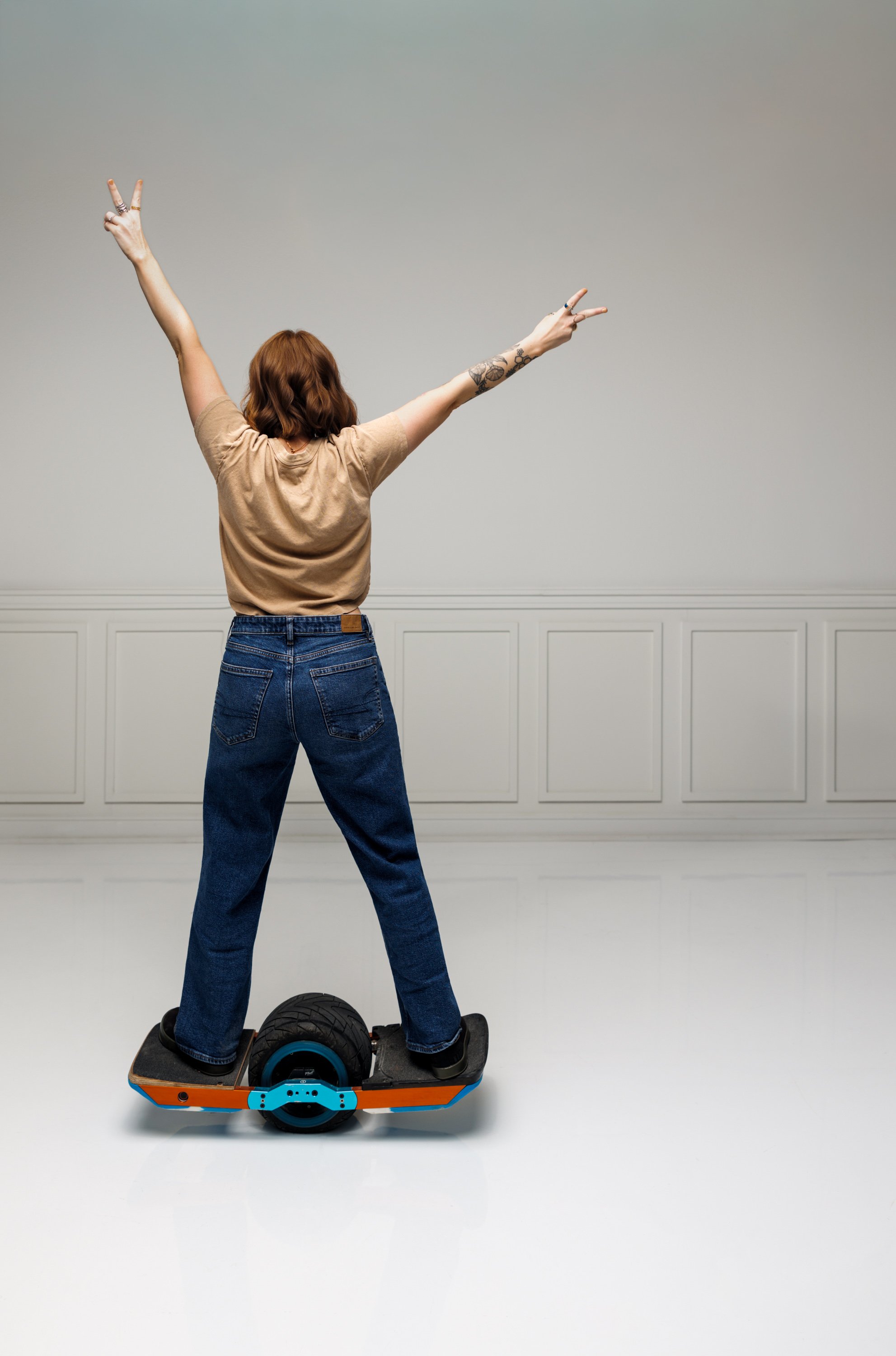 Tiffany Thomas with red hair and tattoos on their arms standing on a hoverboard with arms raised in a V shape, facing away in a plain, light-colored room.