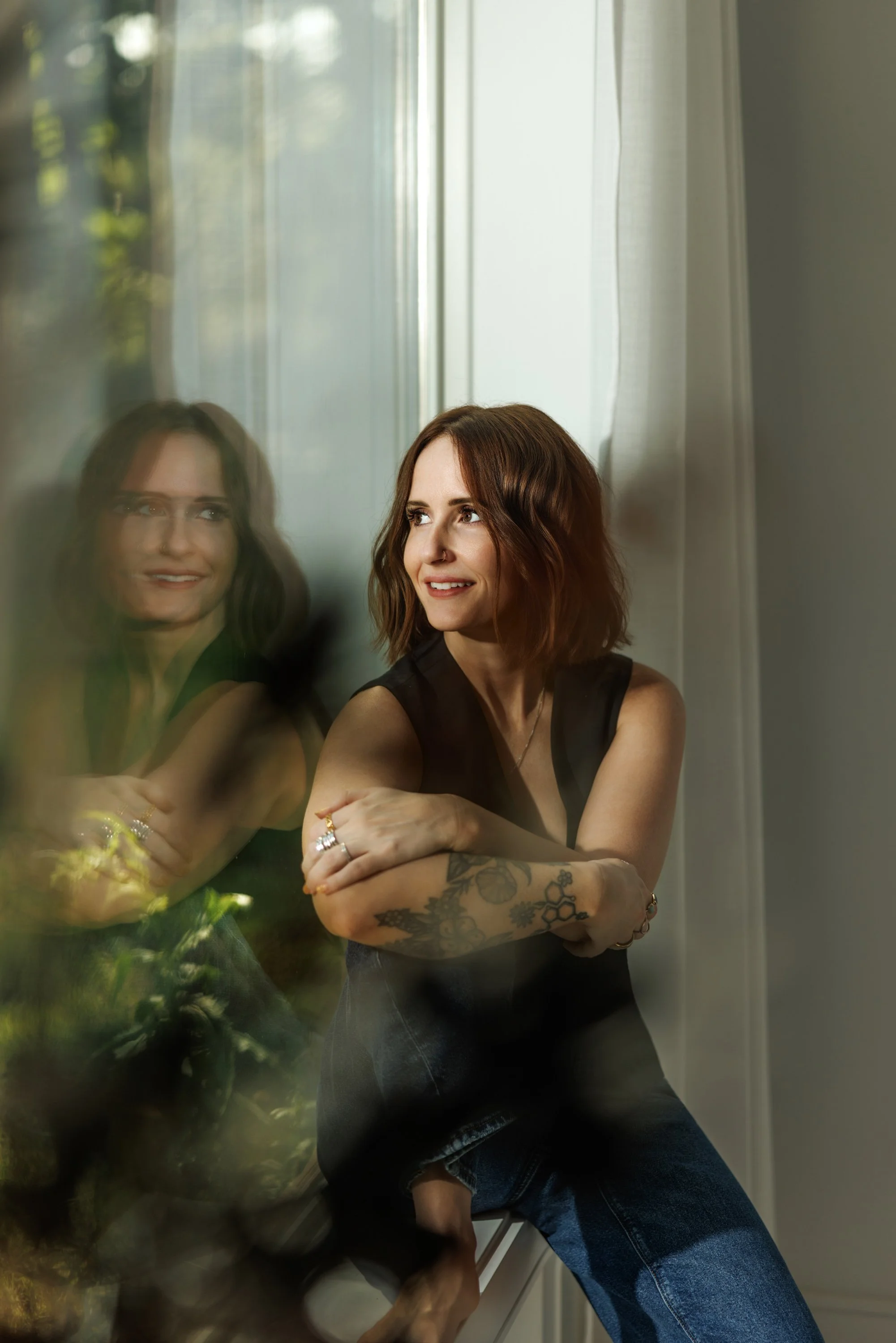 Tiffany Thomas with shoulder-length reddish-brown hair and tattoos on her arms looks to her right, with her reflection visible in a reflective surface, possibly a mirror, in front of her.