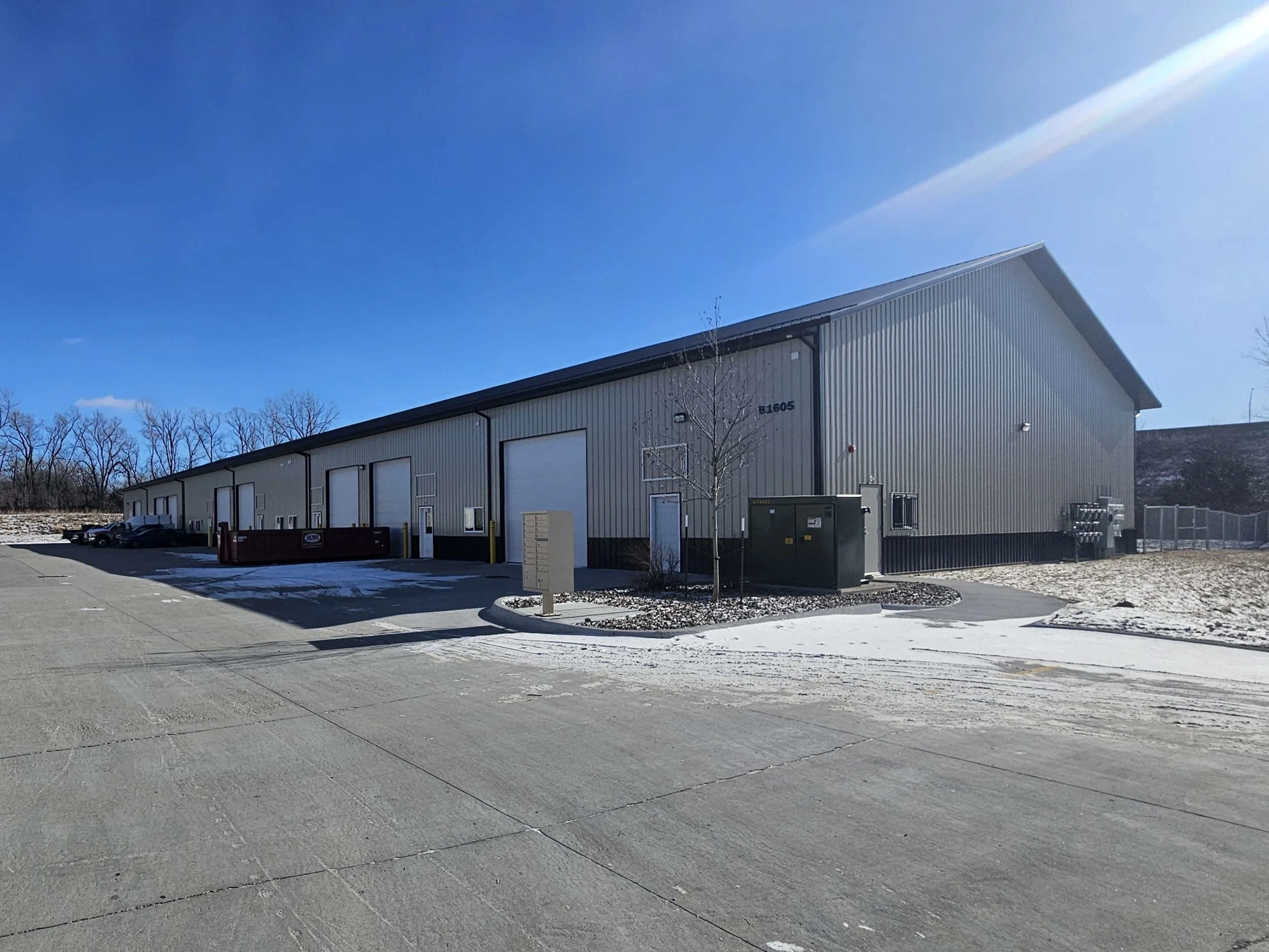 Large industrial warehouse with multiple garage doors, parked trucks, and clear blue sky with bright sunlight.