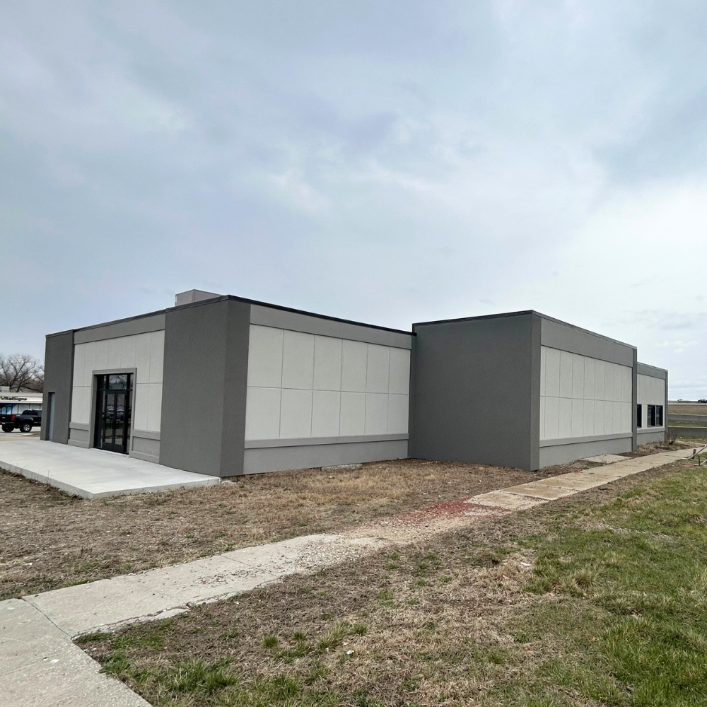 A modern, single-story commercial building with gray and white exterior walls, large windows, and a glass door entrance, surrounded by a mostly dirt lot with some grass and concrete walkways under an overcast sky.