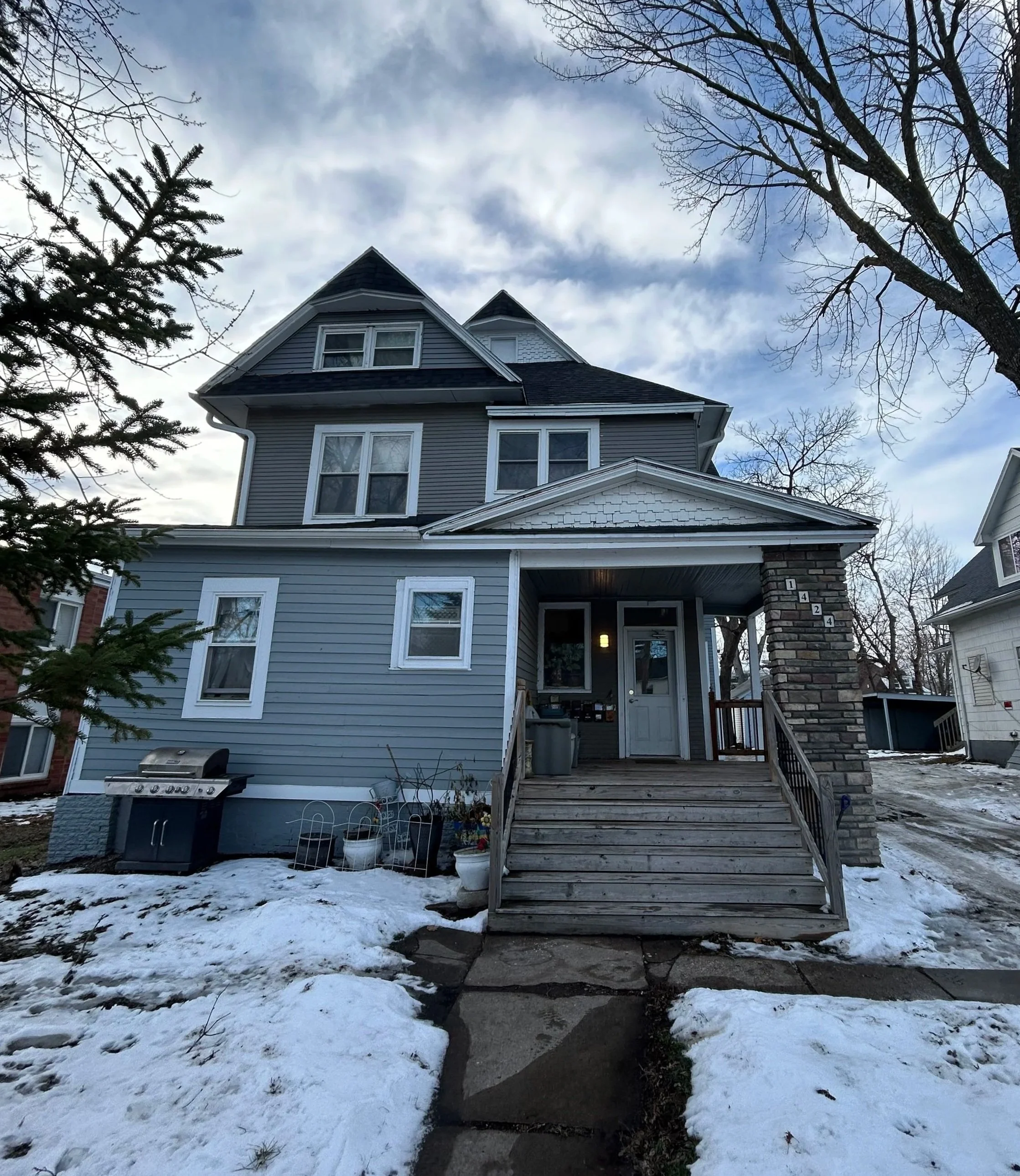 A multi-story gray house with a porch, steps, and a front door, surrounded by snow on the ground and leafless trees in the background.