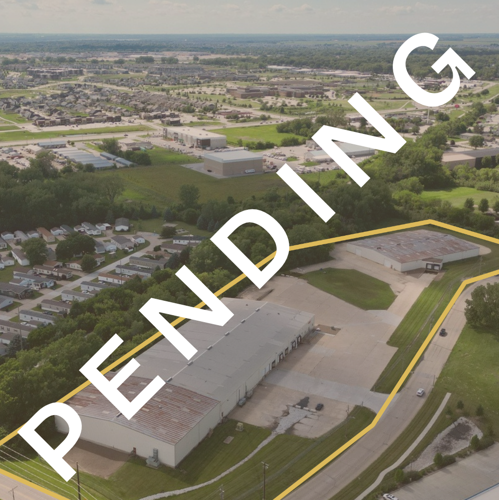 Aerial view of a commercial and industrial area with large warehouse buildings, parking lots, a road, and surrounding greenery and residential neighborhoods.
