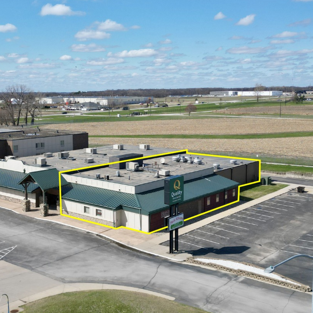 An aerial view of a commercial building with a green roof, marked with a yellow outline. The building has a sign that reads 'Quality' with additional smaller signs below. Adjacent to the building is a parking lot with no cars parked, and the surrounding area includes fields and a few trees under a partly cloudy sky.
