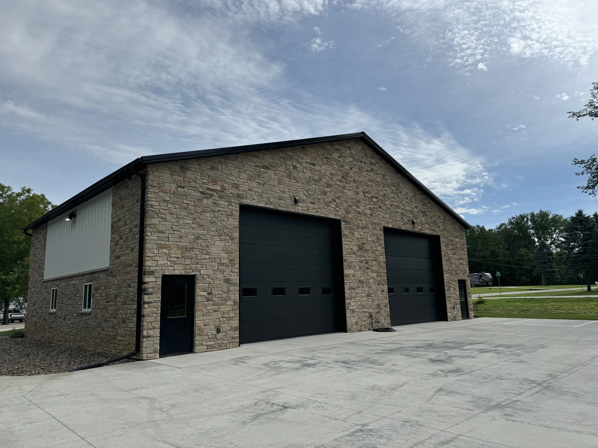 A large modern building with a brick facade and two black garage doors, situated on a concrete lot with surrounding greenery and a partly cloudy sky overhead.