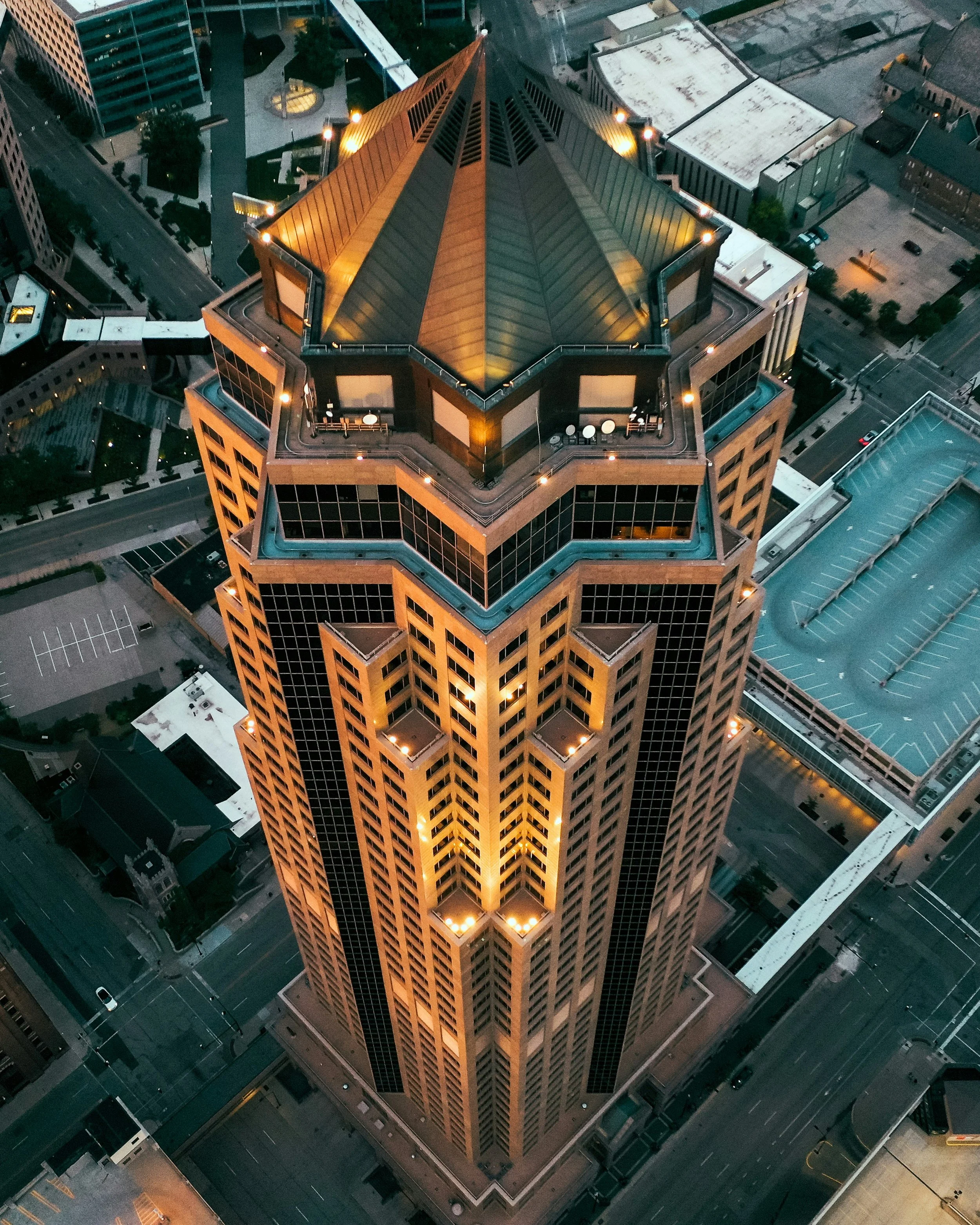 Aerial view of a tall, illuminated skyscraper in a city at dusk, showing its unique angular roof and grid-patterned windows.
