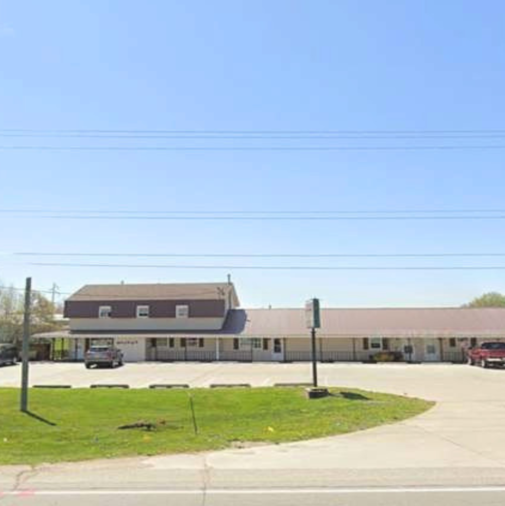 A single-story building with a parking lot in front and a grassy area. The building has a second floor section with a different color and style. The sky is clear and blue.