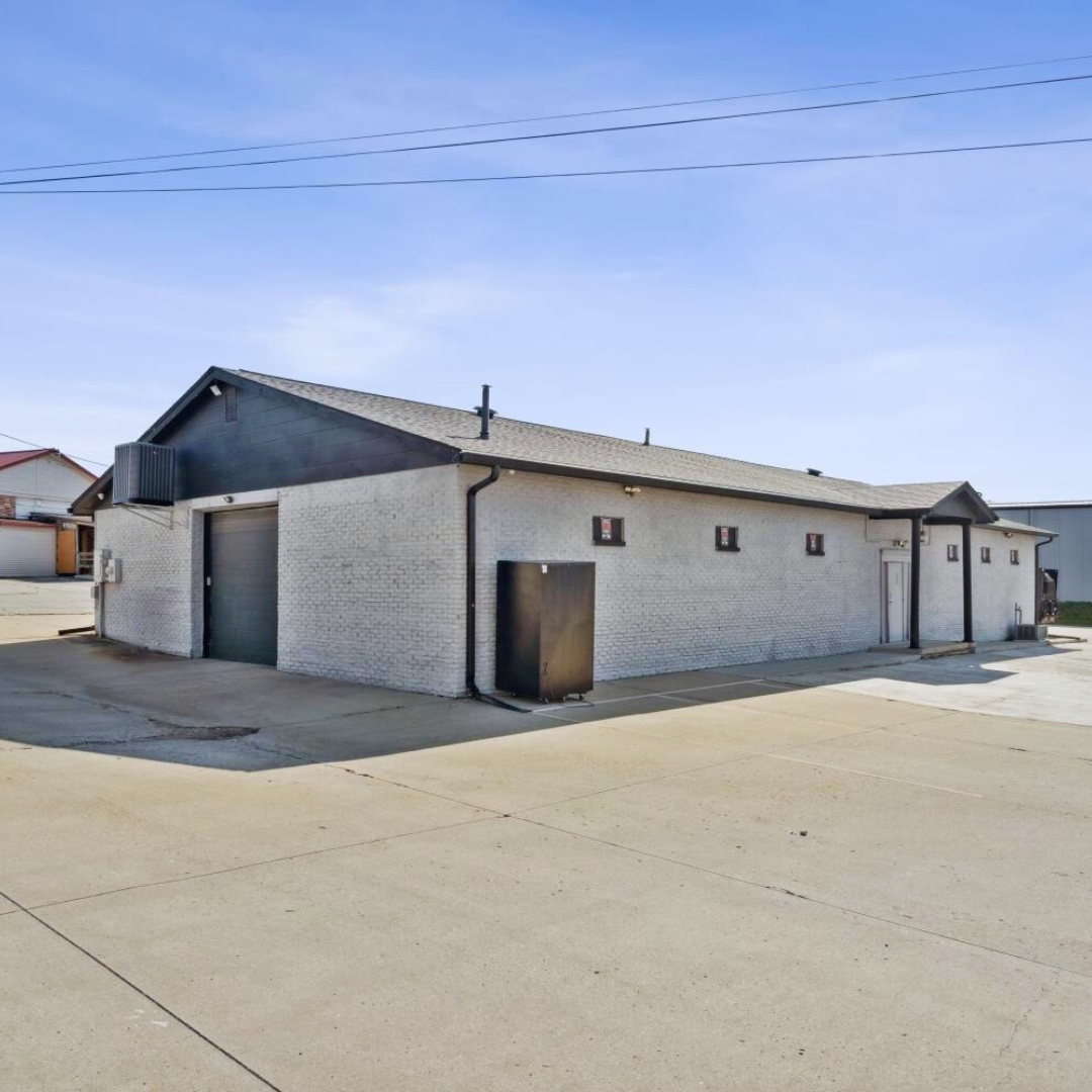 A single-story white brick building with a dark sloped roof, several small windows, and large doors on a concrete lot under a blue sky.
