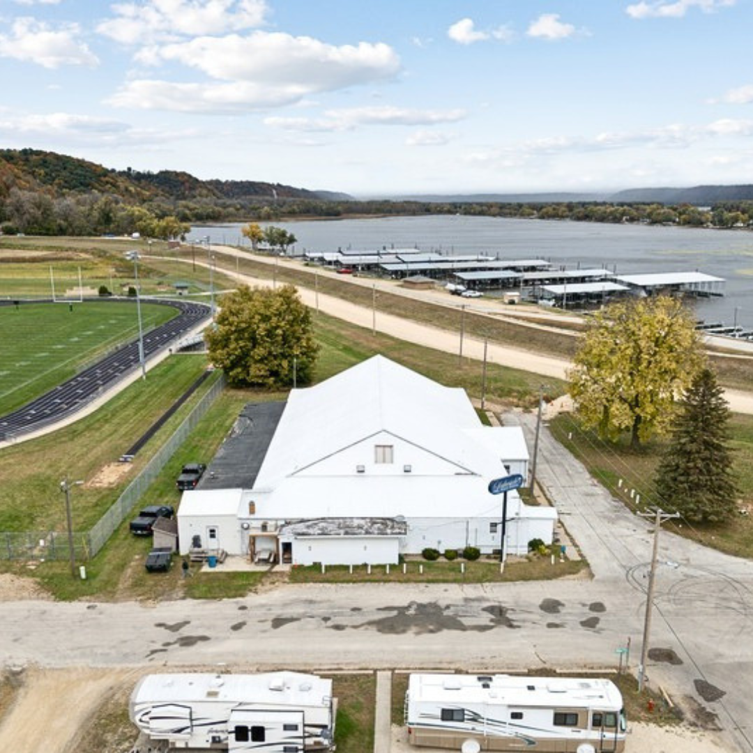 Aerial view of a white building with a football field on the left, a waterfront with boat docks in the background, and trees around the property.