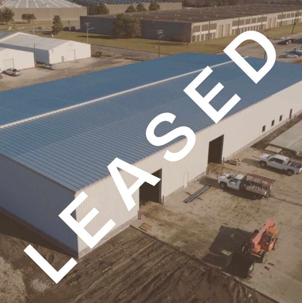 An aerial view of a large industrial warehouse with a blue metal roof, multiple vehicle entrances, and surrounding construction area with trucks and equipment.