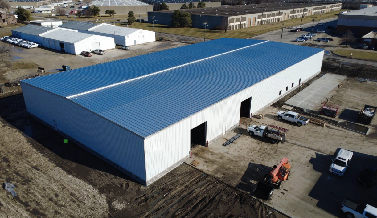 An aerial view of a large industrial warehouse with a blue metal roof, multiple vehicle entrances, and surrounding construction area with trucks and equipment.
