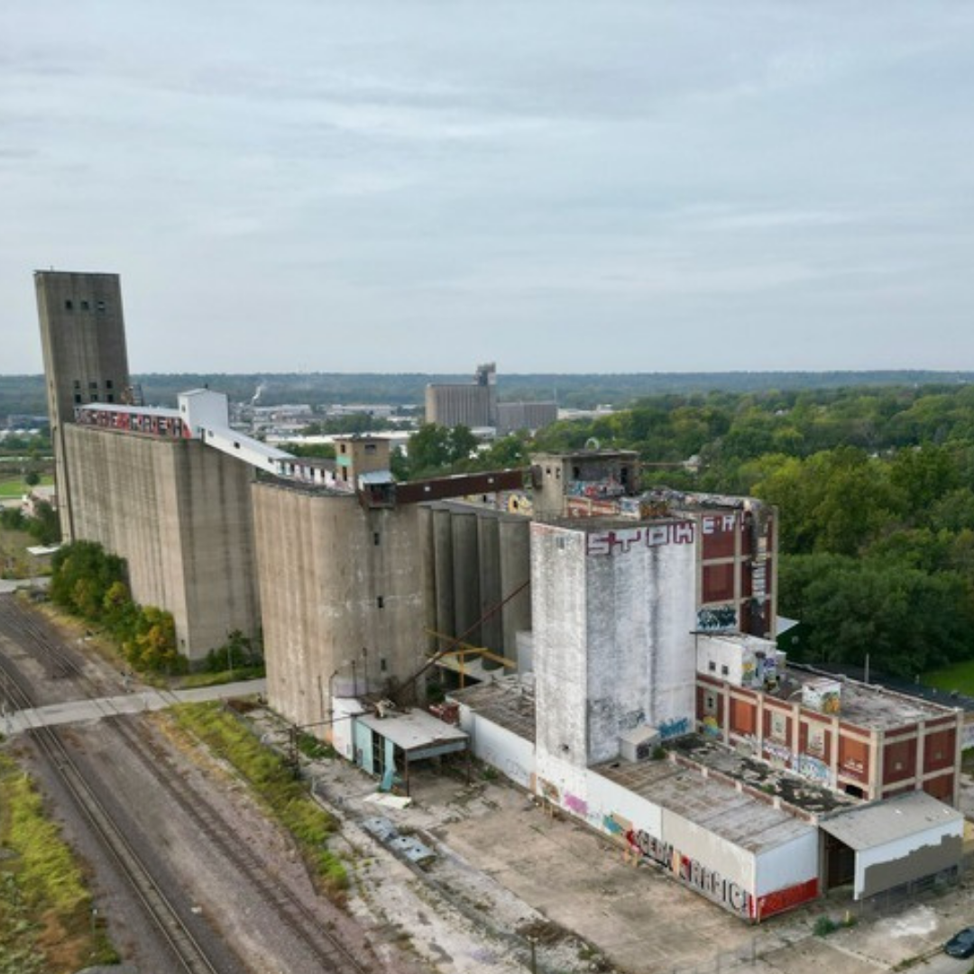 An abandoned, graffiti-covered building with tall concrete silos, situated near train tracks in a green, urban area.