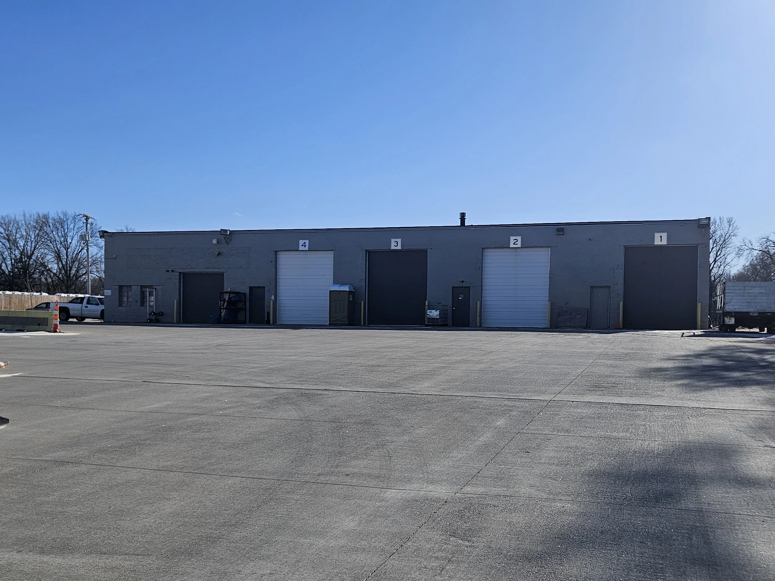 Large industrial warehouse with five loading dock doors numbered 4, 3, 2, 1, and an additional unnumbered door, situated in an expansive parking lot on a clear, sunny day.