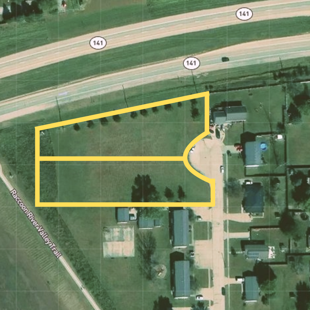 Aerial view of a residential area with a yellow outline marking a specific property, showing houses, driveways, green lawns, and trees, with a road labeled 141 in the background.